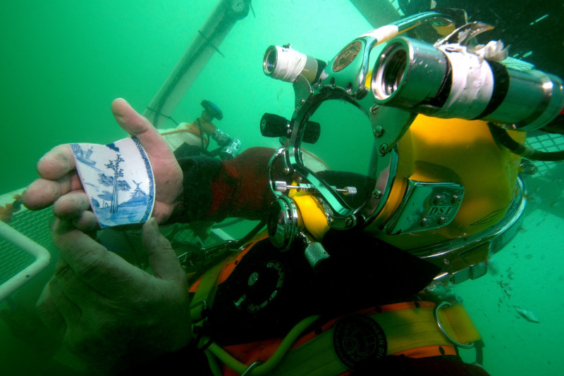 A diver in a diver lift returning to the surface shows a piece of porcelain from the wreck of the VOC ship Rooswijk. Photo #Rooswijk1740 project